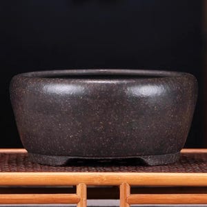 May include: A dark brown ceramic bonsai pot with a rounded shape and three small feet. The pot is sitting on a woven bamboo mat.
