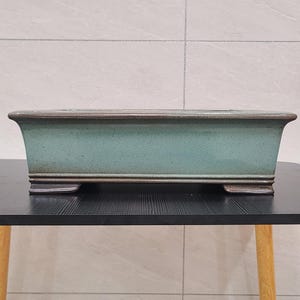 Puede incluir: Una maceta rectangular de cerámica para bonsái con un esmalte verde claro y un borde marrón oscuro. La maceta tiene cuatro pequeños pies y está sentada sobre una mesa negra.