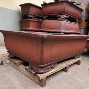 May include: A large, rectangular, reddish-brown ceramic bonsai pot with a wide rim, smooth side panels, and four ornate, curved feet. The pot is positioned on a wooden pallet, with several similar stacked pots visible in the background.