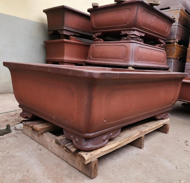 May include: A large, rectangular, reddish-brown ceramic bonsai pot with a wide rim, smooth side panels, and four ornate, curved feet. The pot is positioned on a wooden pallet, with several similar stacked pots visible in the background.