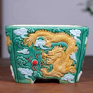 May include: A square green ceramic planter with a raised design of a golden dragon with blue clouds and a red circle.