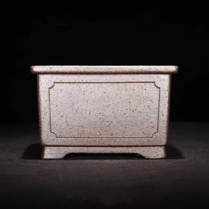 May include: A rectangular brown ceramic planter with a textured surface and a decorative border around the center. The planter has a slightly raised base.