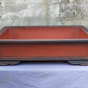Puede incluir: Una maceta rectangular de bonsái roja con un borde marrón oscuro y tres patas decorativas. La maceta está hecha de cerámica y tiene un acabado liso y brillante.