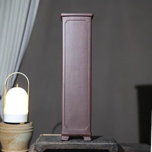 May include: A tall, rectangular, dark reddish-brown cabinet with a textured surface. The cabinet has a decorative frame around the front panel and sits on four small feet. The cabinet is displayed on a dark, ornate wooden surface.