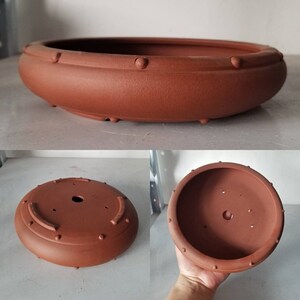 K&ouml;nnte beinhalten: Ein runder, flacher Bonsai-Topf aus Terrakotta mit einem erh&ouml;hten Rand. Der Topf hat eine strukturierte Oberfl&auml;che und ein Abzugsloch im Boden.