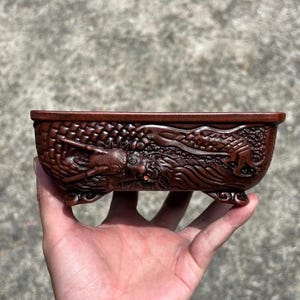 May include: A rectangular, brown, carved planter with a dragon design. The planter is held in a hand and features intricate details of a dragon along the sides. The planter is approximately 8 inches long.