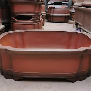 Puede incluir: Una jardinera rectangular grande, de color terracota, con un borde festoneado y una base marrón oscuro. La jardinera tiene una superficie lisa y está diseñada para uso en exteriores. Otras jardineras similares son visibles en el fondo.