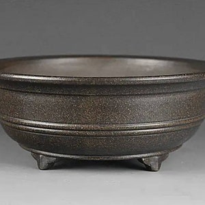 May include: A dark brown ceramic bonsai pot with three short legs. The pot has a textured surface and a slightly rounded shape.