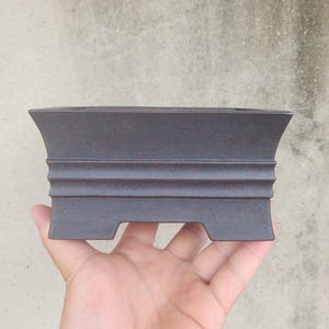 May include: A rectangular bonsai pot with a dark brown finish and a textured surface. The pot has a flared top and a decorative, ridged design along the sides. The pot is designed for planting small trees and plants.