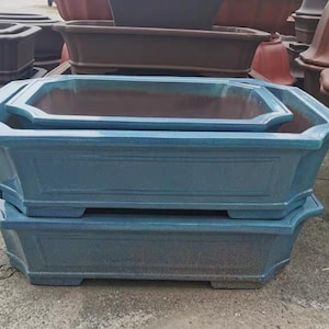 Puede incluir: Pila de tres macetas rectangulares azul-verdosas para bonsái con acabado brillante. Las macetas tienen un diseño clásico con un borde ligeramente ensanchado y una base rectangular. Otras macetas marrones y negras están en el fondo.
