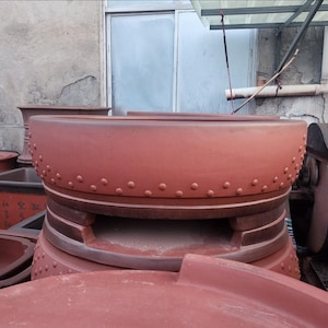 May include: A collection of terracotta-colored ceramic planters. The large, round planter in the foreground has a wide rim with evenly spaced, raised dots. The base of the planter has a stepped design. Other planters of various shapes are visible in the background.