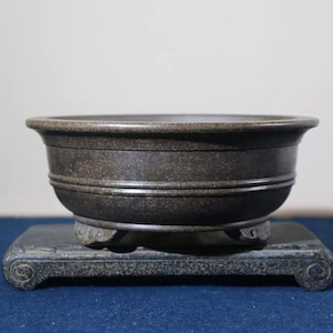 Può includere: Un vaso per bonsai in ceramica marrone scuro con tre gambe e una superficie testurizzata. Il vaso è posto su un supporto in legno nero.