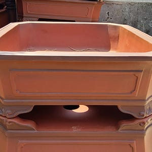 Pode incluir: Um vaso retangular de cor terracota com um topo octogonal. O vaso tem pain&eacute;is decorativos e uma base detalhada. V&aacute;rios vasos semelhantes est&atilde;o empilhados no fundo, sugerindo um tema de jardim ou exterior.