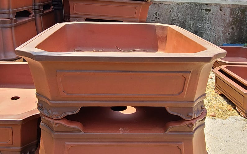 Pode incluir: Um vaso retangular de cor terracota com um topo octogonal. O vaso tem pain&eacute;is decorativos e uma base detalhada. V&aacute;rios vasos semelhantes est&atilde;o empilhados no fundo, sugerindo um tema de jardim ou exterior.