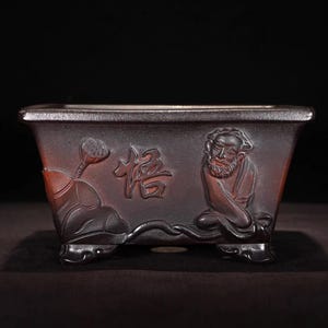 Puede incluir: Una maceta rectangular de cerámica marrón oscuro para bonsái con un relieve tallado de una figura barbuda, una flor de loto y el carácter chino 悟. El recipiente tiene un borde acampanado, acabado liso y cuatro pies pequeños, ornamentados y curvos.