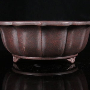 Pode incluir: Um vaso de bonsai de cerâmica marrom escuro com uma borda festonada e três pequenos pés. O vaso tem uma superfície texturizada e uma forma ligeiramente arredondada.