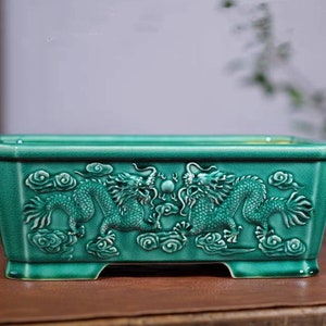May include: A rectangular, teal-colored ceramic planter with a glossy finish. The planter features a detailed relief of two dragons facing each other, set against a backdrop of clouds. The design is symmetrical, with the dragons flanking a central orb.