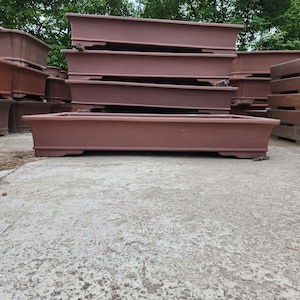 May include: A stack of rectangular, reddish-brown bonsai pots. The pots have a low profile, with a slightly flared rim and a raised base. The pots are made of a textured material and are designed for growing bonsai trees.