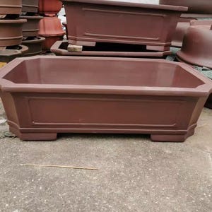 May include: A rectangular brown ceramic planter with a decorative rim and a raised rectangular design on the front. The planter is sitting on a concrete surface.