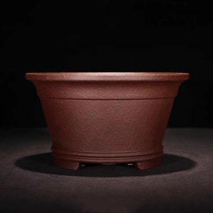 Puede incluir: Una maceta de bonsái de color rojo oscuro, de forma ovalada, con una superficie texturizada y un borde ligeramente acampanado. La maceta tiene tres pequeños pies en la parte inferior.