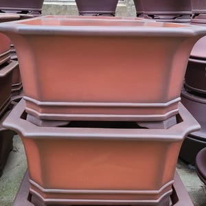 Peut inclure: Deux pots en terre cuite, l'un empilé sur l'autre. Les pots sont non émaillés et de couleur brun rougeâtre. Les pots sont rectangulaires avec des coins arrondis et ont une lèvre autour du bord supérieur.