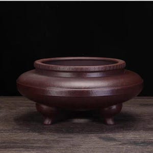 May include: A maroon-colored ceramic incense burner with a wide, rounded body and a decorative band around the rim. The burner rests on three small, rounded feet. The background is a dark, solid color.