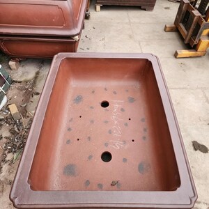 May include: A large, rectangular, unglazed terracotta bonsai pot with a wide, flat rim. The interior base features two large drainage holes, four smaller drainage holes, and handwritten white text reading 41.7x56x20.1cm.
