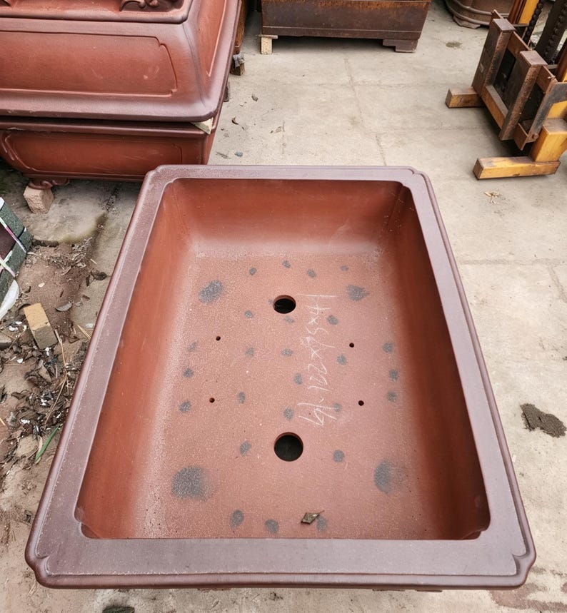 May include: A large, rectangular, unglazed terracotta bonsai pot with a wide, flat rim. The interior base features two large drainage holes, four smaller drainage holes, and handwritten white text reading 41.7x56x20.1cm.
