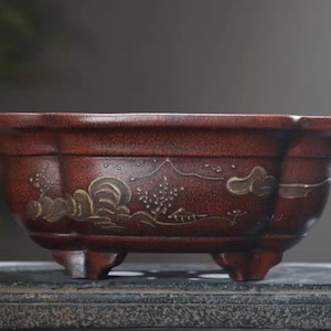 May include: A red ceramic bonsai pot with a decorative design of mountains and clouds in gold. The pot has four legs and a rounded shape.
