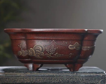 May include: A red ceramic bonsai pot with a decorative design of mountains and clouds in gold. The pot has four legs and a rounded shape.