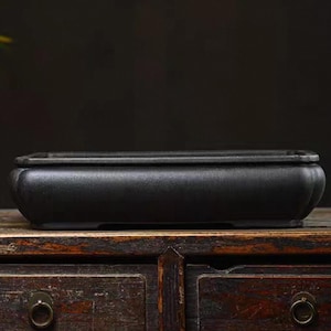 Puede incluir: Una maceta rectangular de bonsai de cerámica negra con bordes redondeados. La maceta está sobre una superficie de madera.