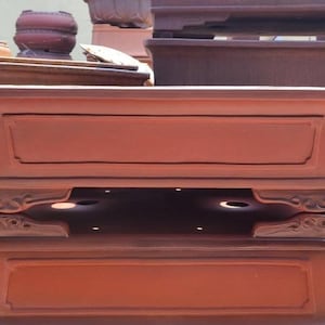 May include: A set of three terracotta plant pots with intricate detailing. The pots are stacked on top of each other, with the largest pot at the bottom and the smallest pot at the top. The pots are a reddish-brown color and have a smooth, slightly textured surface.