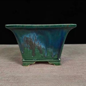 May include: A square, glazed ceramic planter with a flared rim. The planter has a glossy finish with a blend of green and blue hues, with darker streaks. It sits on four small feet, suitable for indoor or outdoor use.