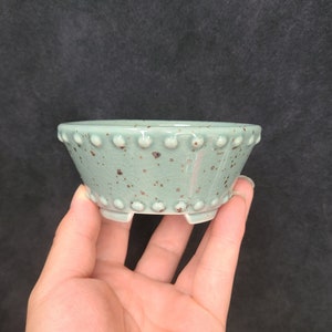 Puede incluir: Una maceta pequeña de cerámica azul claro con un esmalte moteado y un borde elevado. La maceta tiene tres pequeños pies y es perfecta para plantas pequeñas.