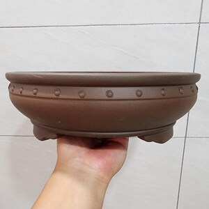 Könnte beinhalten: Braune Bonsaischale aus Keramik mit breitem, flachem Design. Der Topf hat eine abgerundete Form mit einer Reihe kleiner, gleichmäßig verteilter kreisförmiger Vertiefungen am Rand. Er ruht auf drei kleinen Füßen.