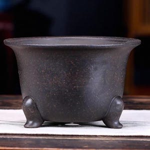 Puede incluir: Una maceta de cerámica gris oscuro con tres patas. La maceta tiene una forma redondeada y una superficie texturizada.