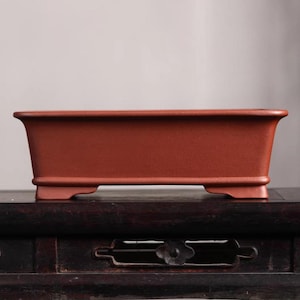 May include: A rectangular terracotta bonsai pot with a slightly flared lip and a flat base. The pot has a reddish-brown color and is sitting on a dark wooden table.