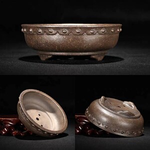 May include: A brown ceramic bonsai pot with a rounded shape and a decorative rim. The pot has three small feet and drainage holes in the bottom.