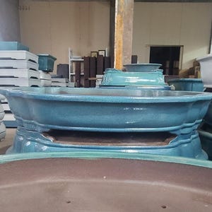 May include: A teal blue ceramic planter with a wide, oval shape and a raised base. The planter has a smooth, glossy finish and is designed for indoor or outdoor use.