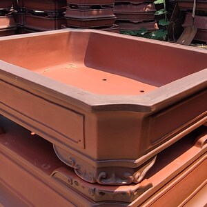 Pode incluir: Uma pilha de vasos retangulares para bonsai na cor terracota. Os vasos t&ecirc;m um design cl&aacute;ssico com molduras decorativas e uma bacia larga e rasa. Os vasos est&atilde;o empilhados uns sobre os outros, sugerindo que est&atilde;o prontos para uso em um jardim.