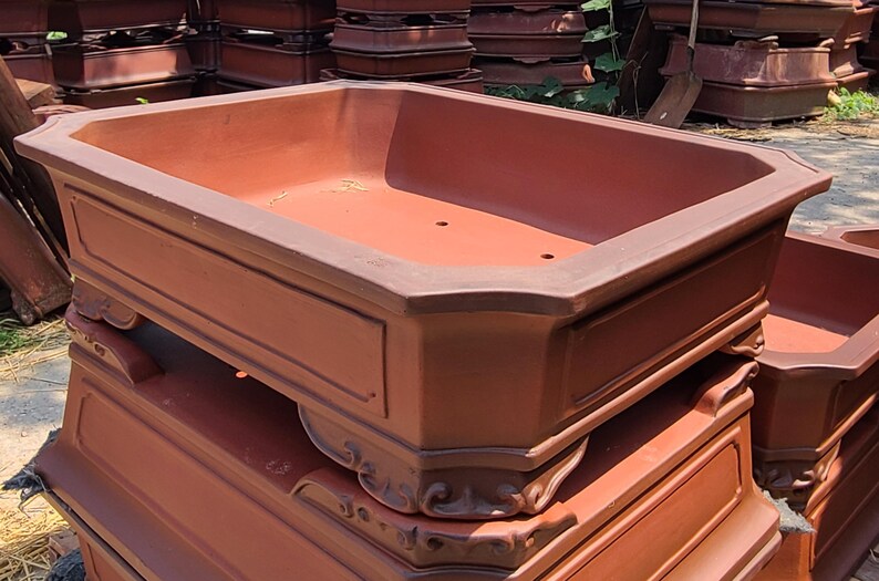 Pode incluir: Uma pilha de vasos retangulares para bonsai na cor terracota. Os vasos t&ecirc;m um design cl&aacute;ssico com molduras decorativas e uma bacia larga e rasa. Os vasos est&atilde;o empilhados uns sobre os outros, sugerindo que est&atilde;o prontos para uso em um jardim.
