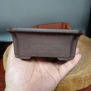 Könnte beinhalten: Ein rechteckiger brauner Keramik-Bonsai-Topf mit leicht ausgestelltem Rand und drei kleinen Füßen an der Unterseite.