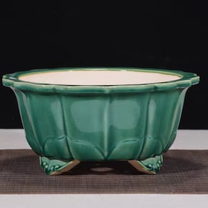 May include: A green ceramic planter with a scalloped rim and three decorative feet. The planter has a glossy finish and a white interior. The sides of the planter are textured with vertical ridges. This decorative planter is suitable for indoor or outdoor use.