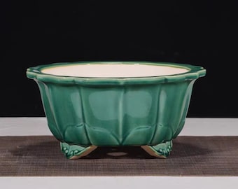 May include: A green ceramic planter with a scalloped rim and three decorative feet. The planter has a glossy finish and a white interior. The sides of the planter are textured with vertical ridges. This decorative planter is suitable for indoor or outdoor use.