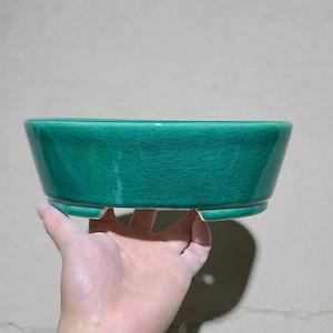 Op de afbeelding: Een groene keramische bonsai pot met een gekrakelde glazuur en een afgeronde ovale vorm. De pot heeft drie kleine voetjes aan de onderkant.