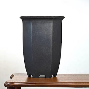 May include: A black, hexagonal ceramic plant pot with a flared rim. The pot has a textured surface and rests on four small feet. It is displayed on a wooden table.