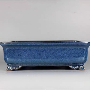 Peut inclure: Un pot à bonsaï carré en céramique bleu avec quatre pieds décoratifs. Le pot a un glaçage bleu moucheté.