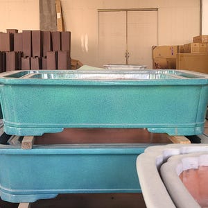 Puede incluir: Dos macetas rectangulares de cerámica turquesa apiladas con una superficie texturizada. Las macetas están vacías y listas para plantar.