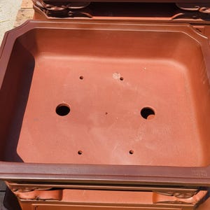 Pode incluir: Um vaso retangular para bonsai, castanho-avermelhado, com uma borda decorativa e orif&iacute;cios de drenagem. O vaso tem um design amplo e aberto, adequado para plantar pequenas &aacute;rvores ou arbustos. O vaso &eacute; feito de um material dur&aacute;vel.