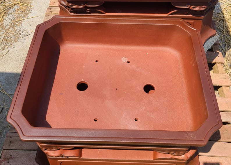 Pode incluir: Um vaso retangular para bonsai, castanho-avermelhado, com uma borda decorativa e orif&iacute;cios de drenagem. O vaso tem um design amplo e aberto, adequado para plantar pequenas &aacute;rvores ou arbustos. O vaso &eacute; feito de um material dur&aacute;vel.